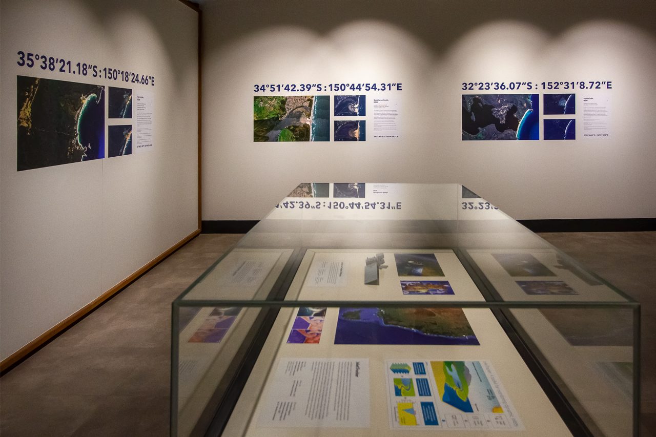 A corner of the Sonus Maris exhibition. In the foreground, a display cabinet with photographs inside. In the background, walls with colourful coastline arial shots.
